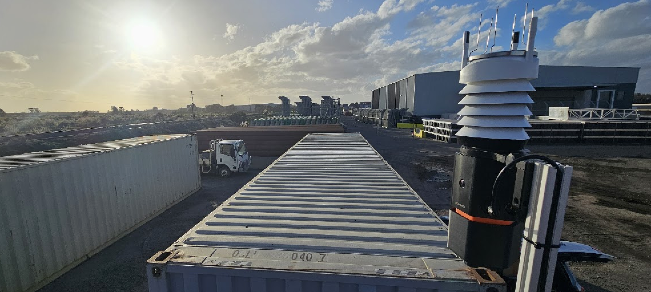 A SiteHive Hexanode Weather device on a pole at the corner of a shipping container, looking out over a container yard with a truck and warehouses on a bright morning